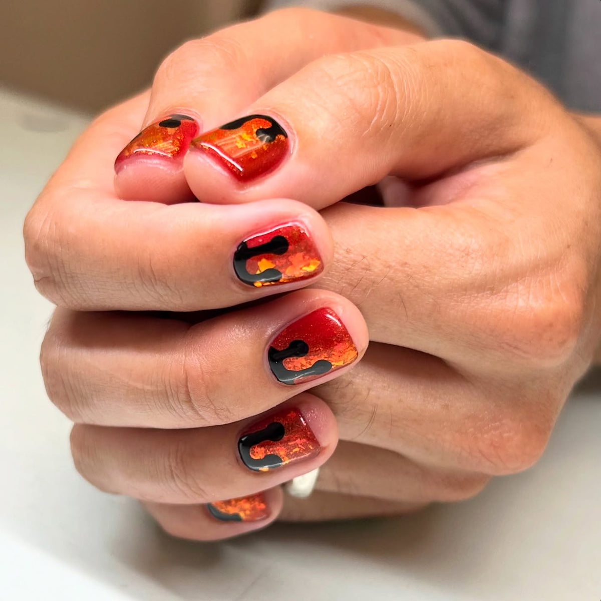 Halloween nail art by nail artist Carrie Huynh featuring Vacation Nail Shop Coral Quartzy Flake, with fiery orange-red foil flakes encapsulated beneath glossy black abstract drip details on short natural nails.