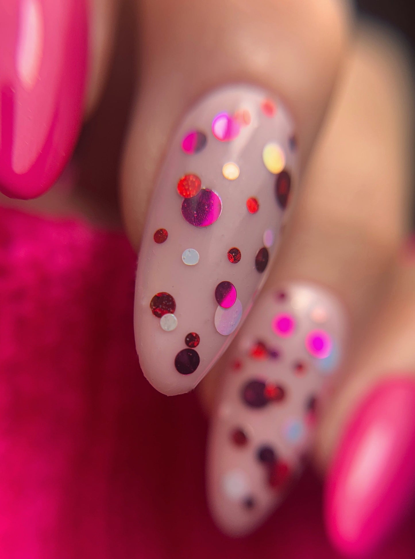 Close-up of a hand with pink nail polish and multicolored confetti dots on a pink background.