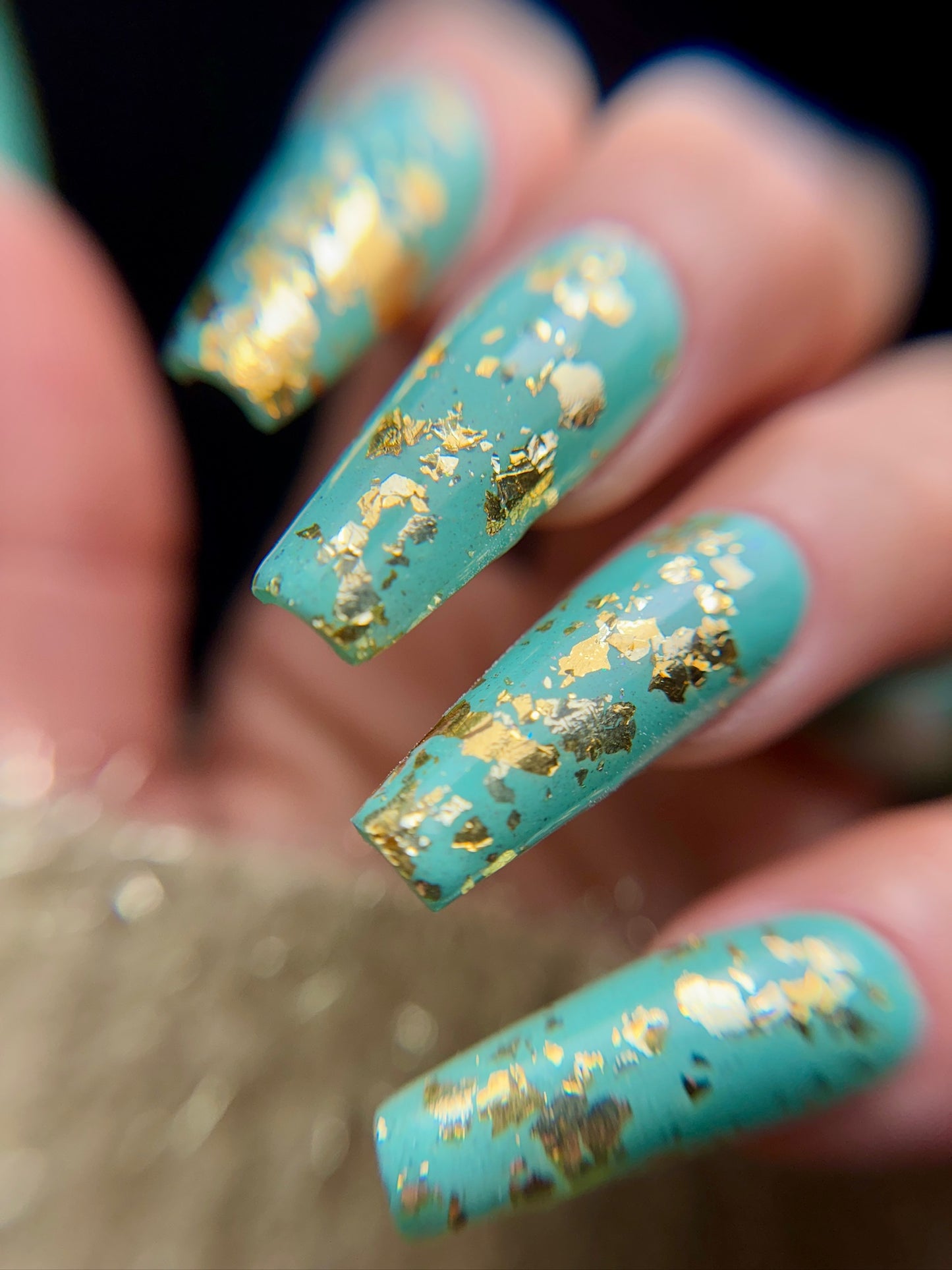 Turquoise nails with gold leaf design against a blurred background