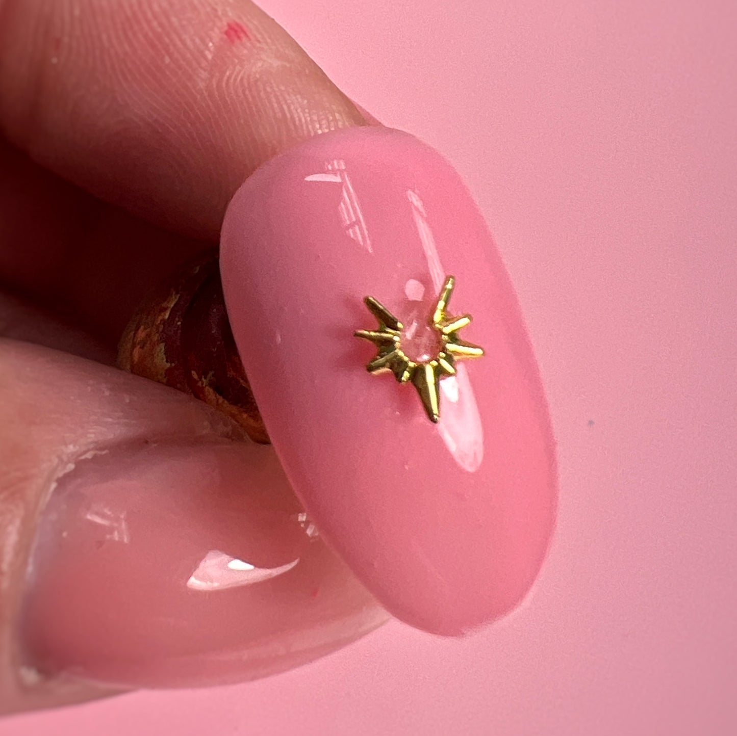 Pink nail with a gold star-shaped design held by a thumb against a pink background
