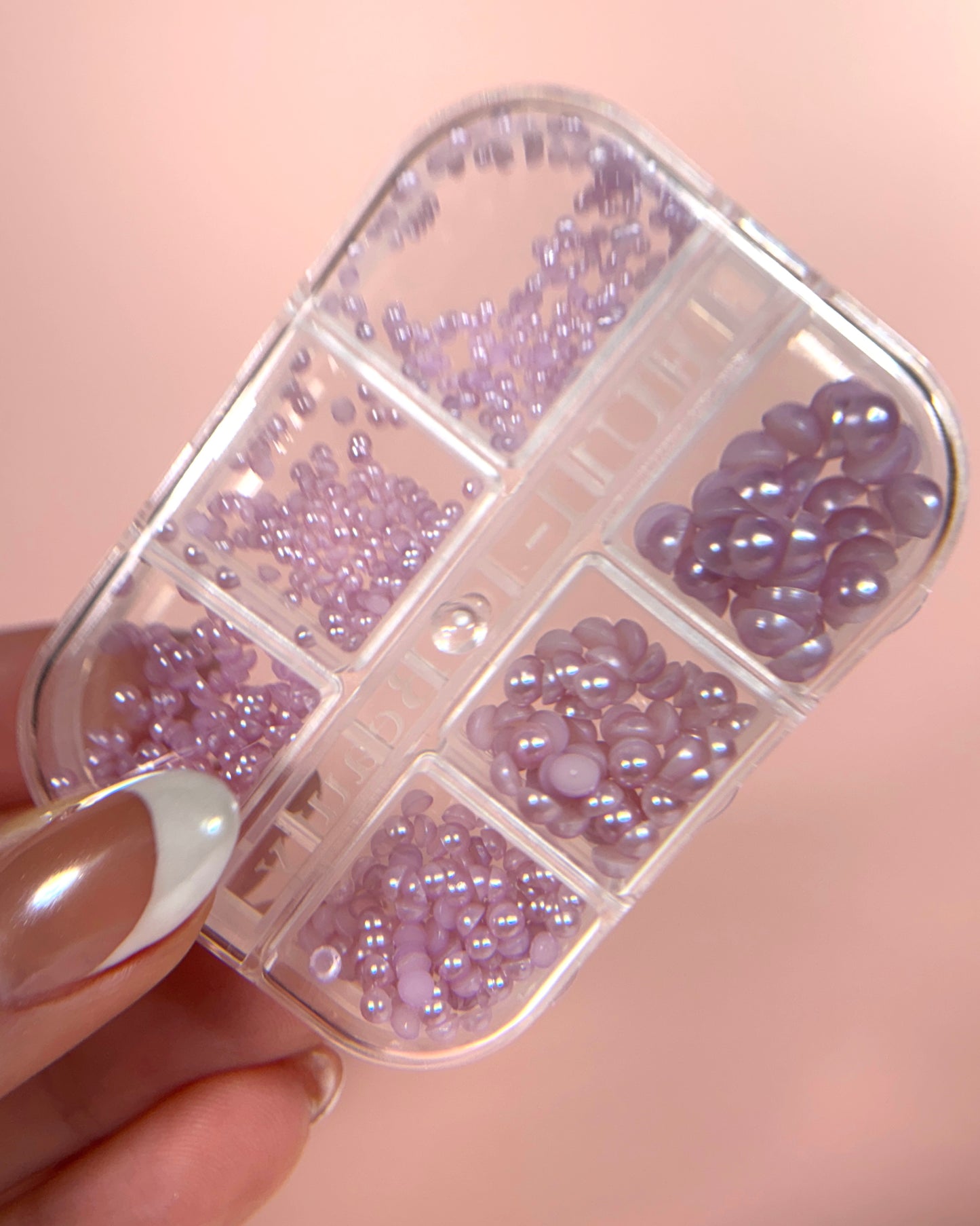 Hand holding transparent container with lavender pearly half-sphere beads on pink background