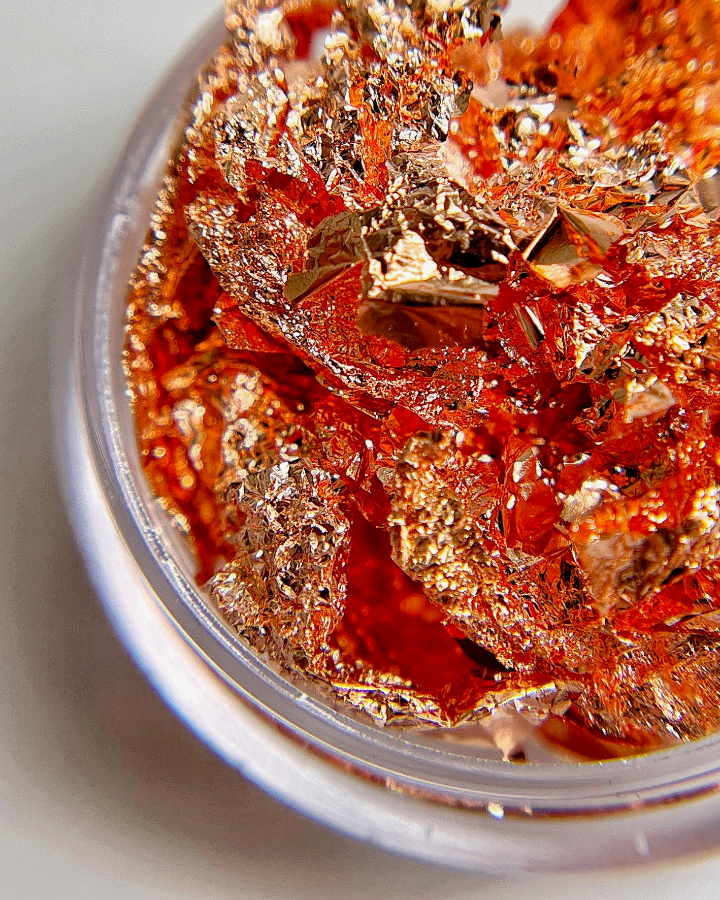 Detailed view of copper colored foil leaf flake in clear jar on white background.