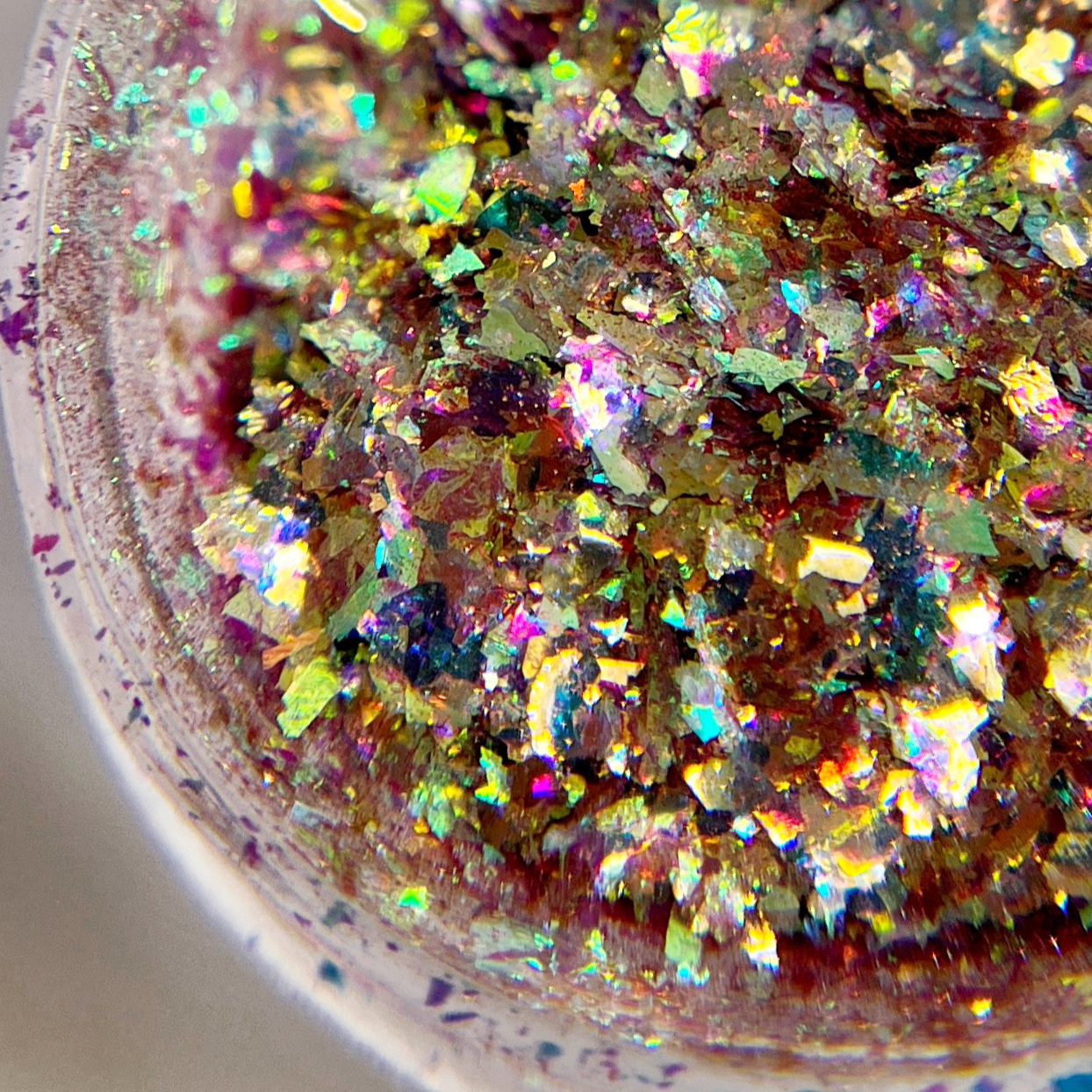 A close-up of nail art flakes with a mix of colors including purple, green, and gold, showcasing their sparkling and multicolored appearance.