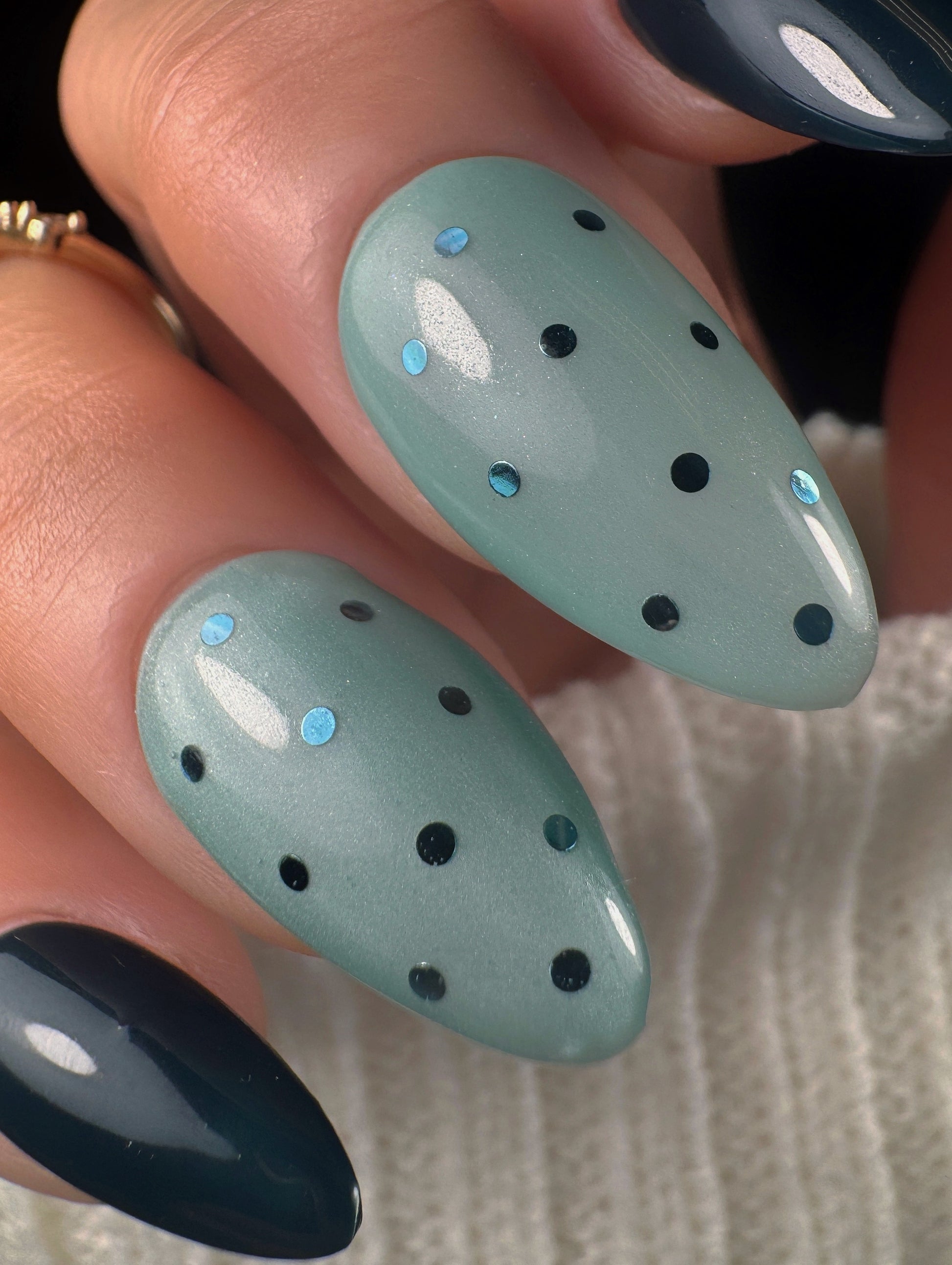 Nails with a light teal base and dark teal polka dots glitter held against a neutral background.