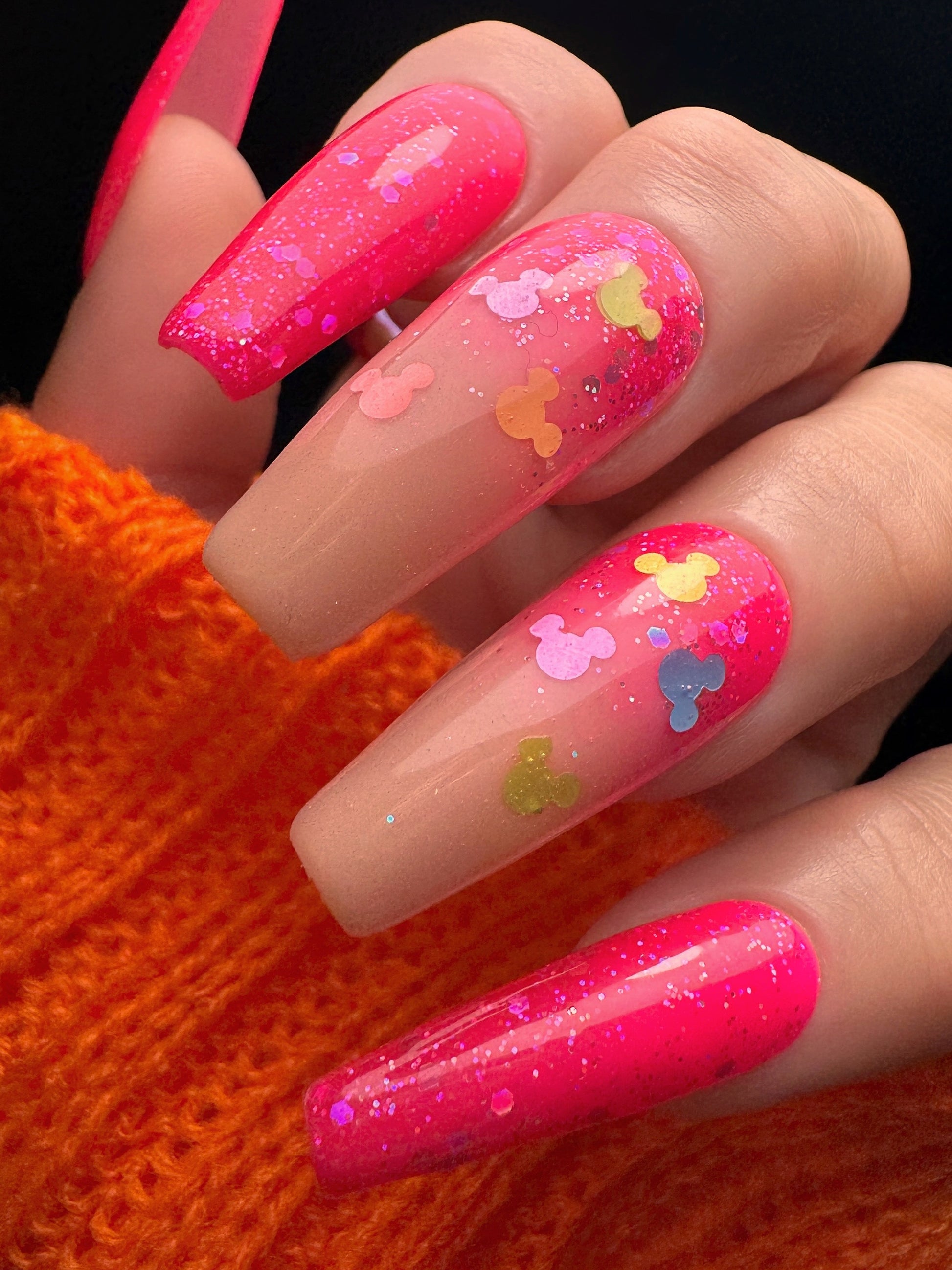 Hand with bright pink nails featuring colorful designs on an orange textured background