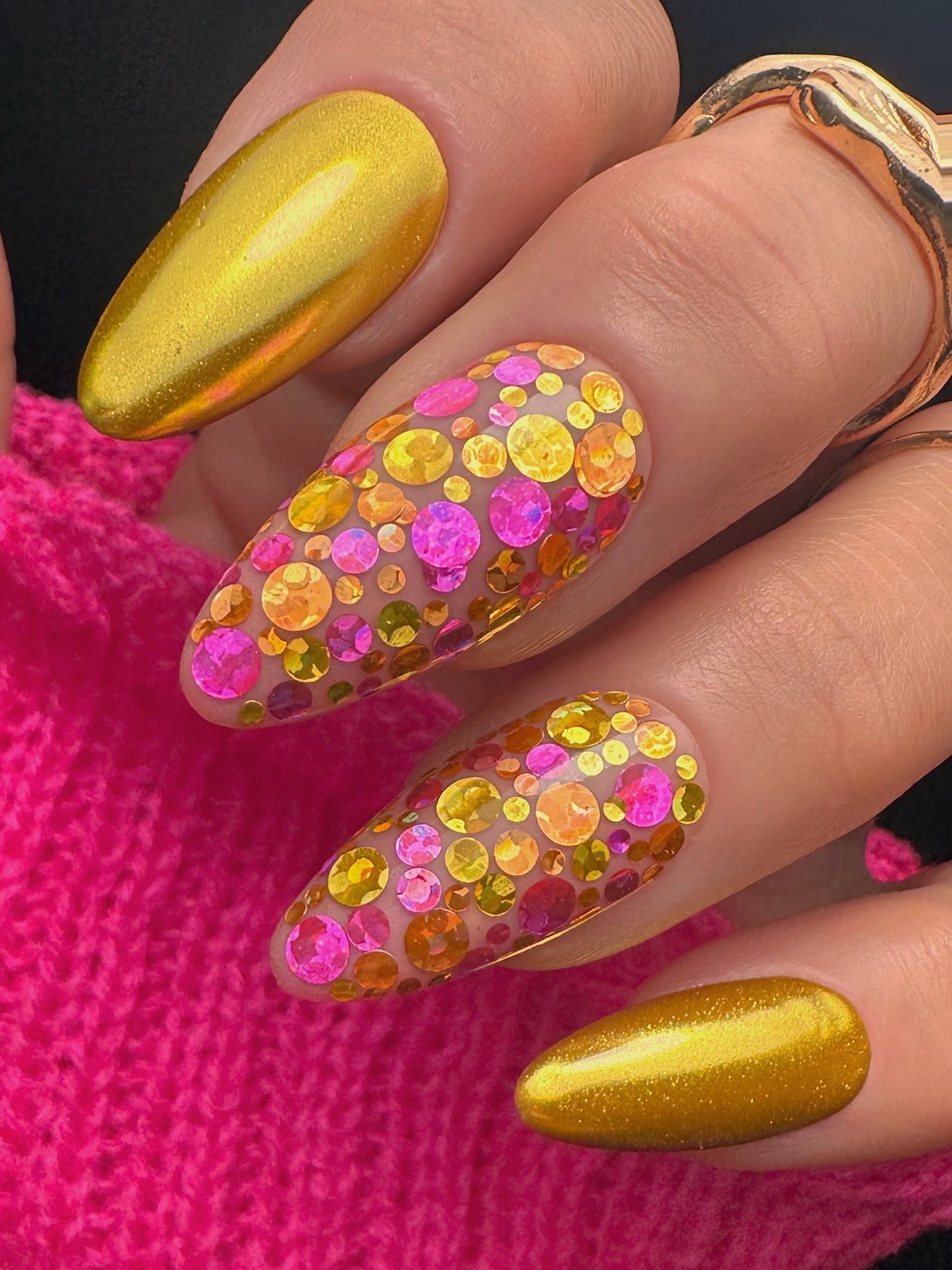 Nails with colorful sequin design and gold polish against a pink background
