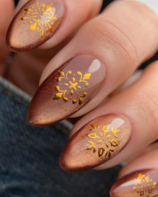 Nails with brown nail polish and gold floral designs on a blurred background