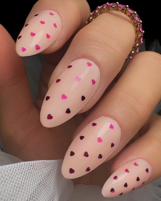 Nails with pink polish and heart-shaped decorations, wearing a gold ring.