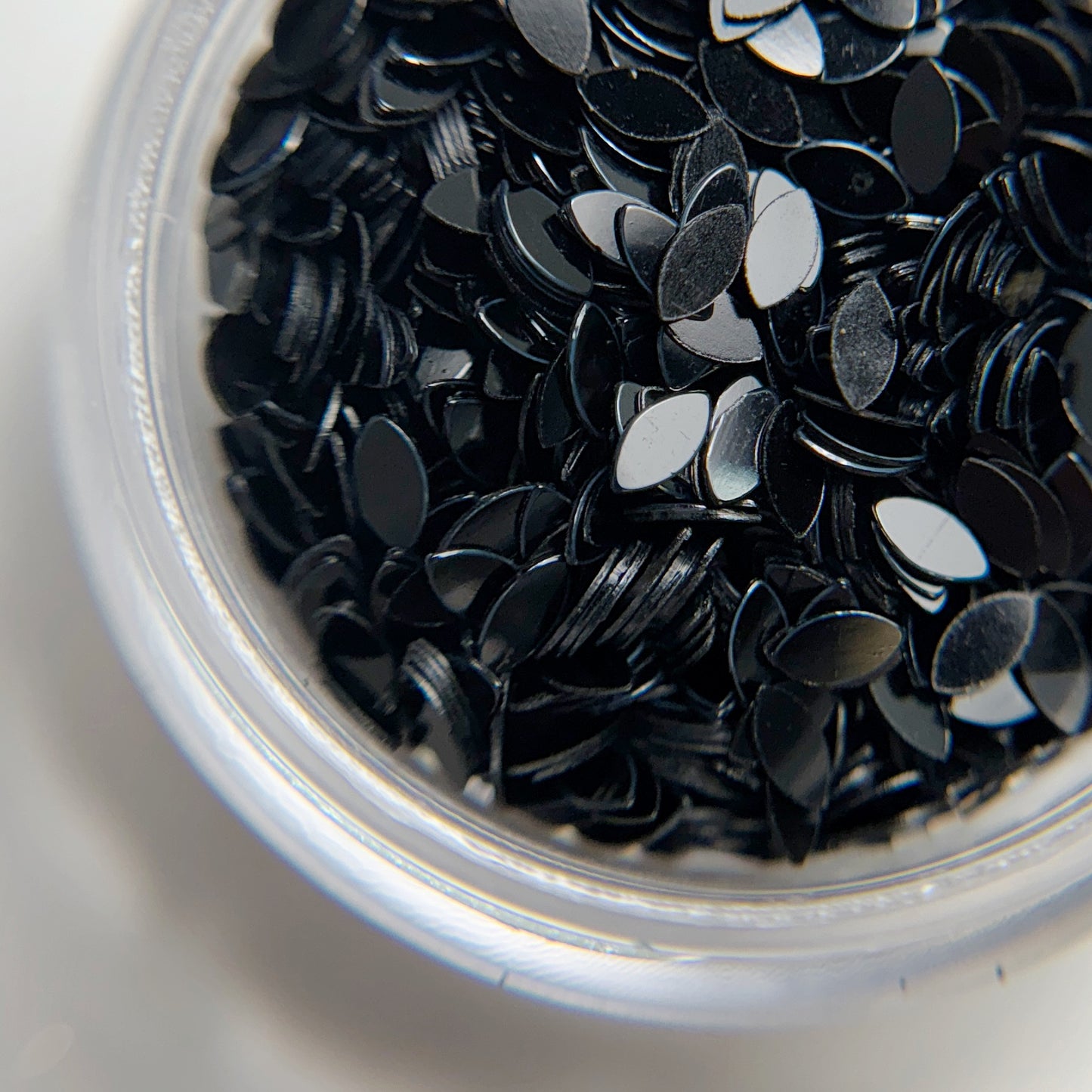 A close-up view of black, dragon scale-shaped glitter contained within a clear plastic jar.