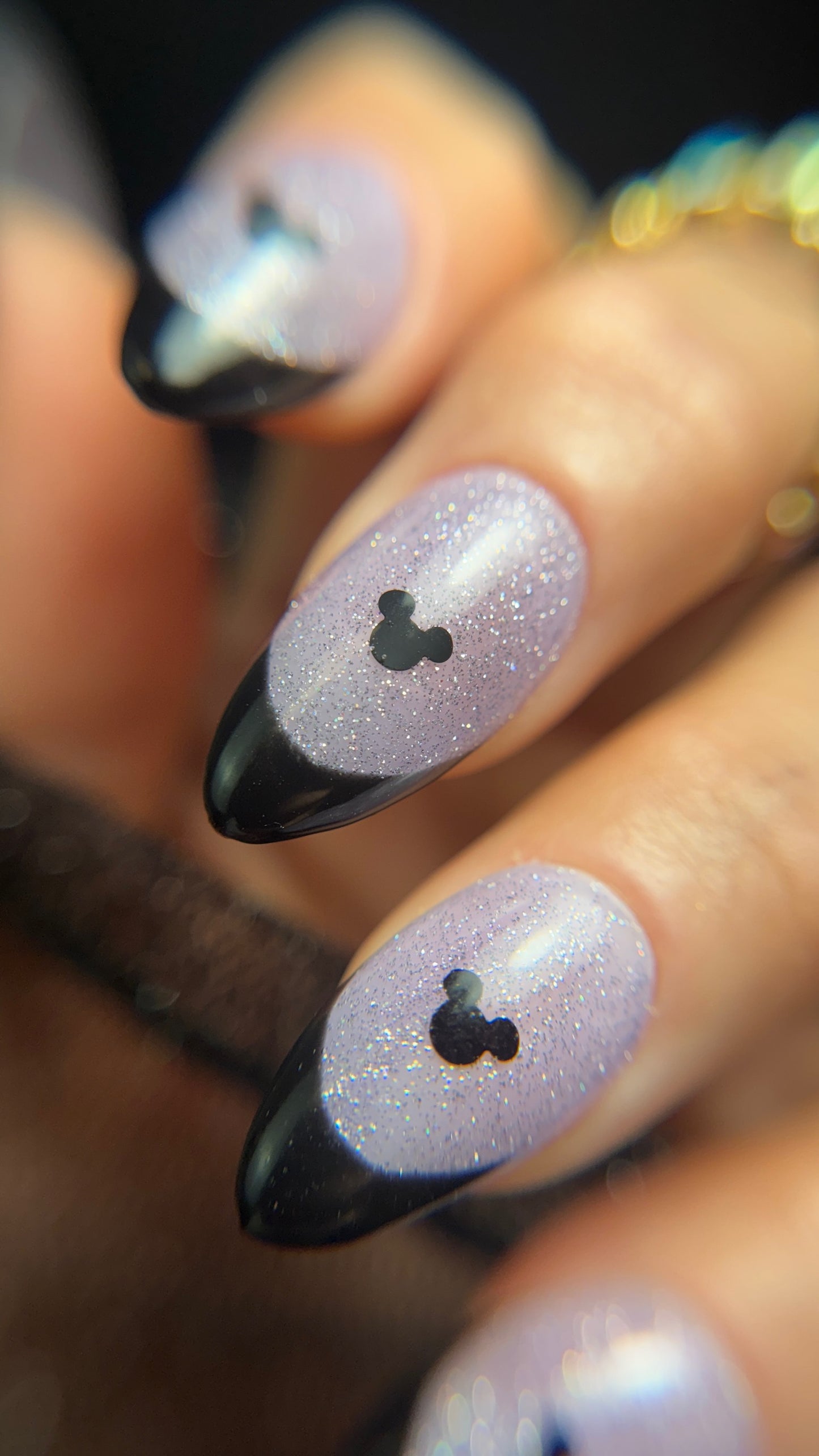 Nails with black and lavender glitter design resembling Mickey Mouse, held by a hand.