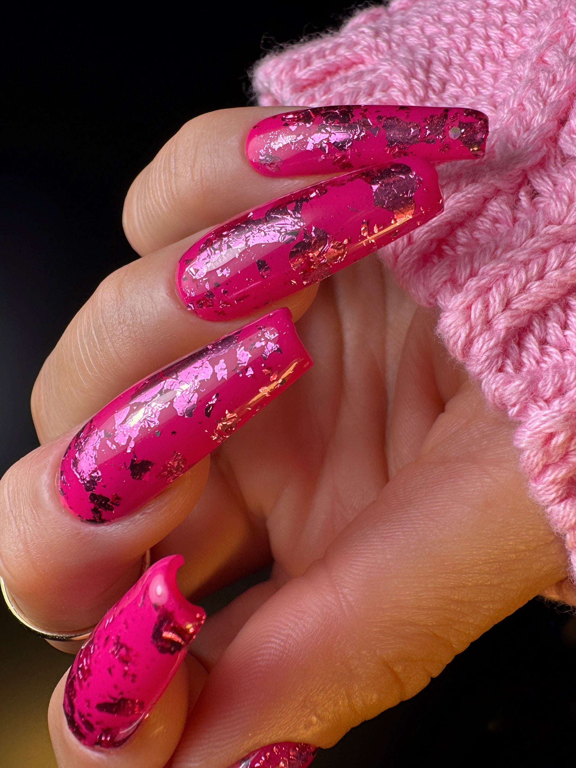 Hand with pink glittery nails wearing a pink knitted sleeve against a black background