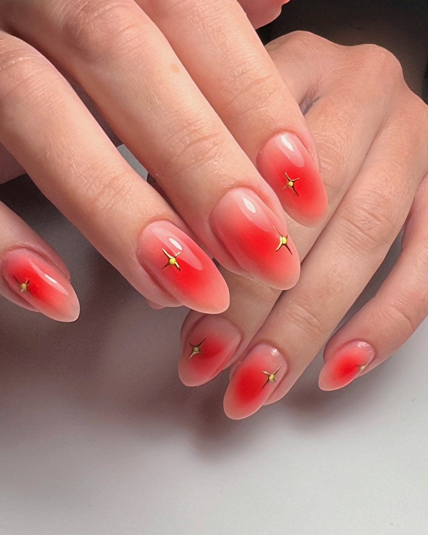 A close-up image of a person's fingernails, which are painted with a red gel polish and adorned with small gold accents.