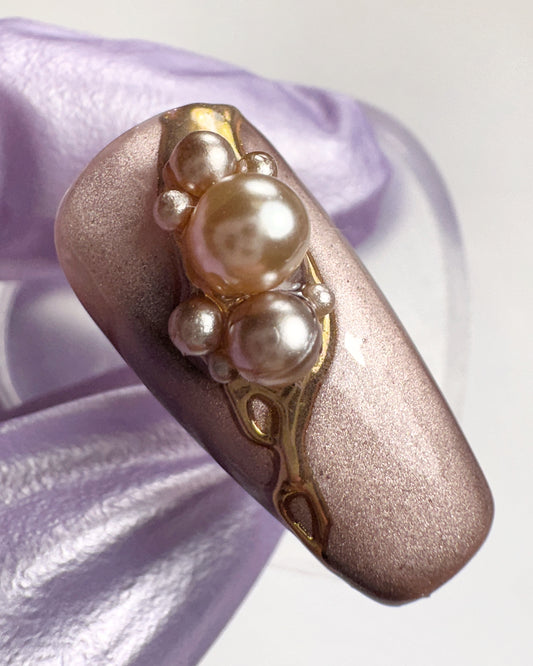Nail art with pearls and gold accents on a pinkish-brown nail against a light purple background
