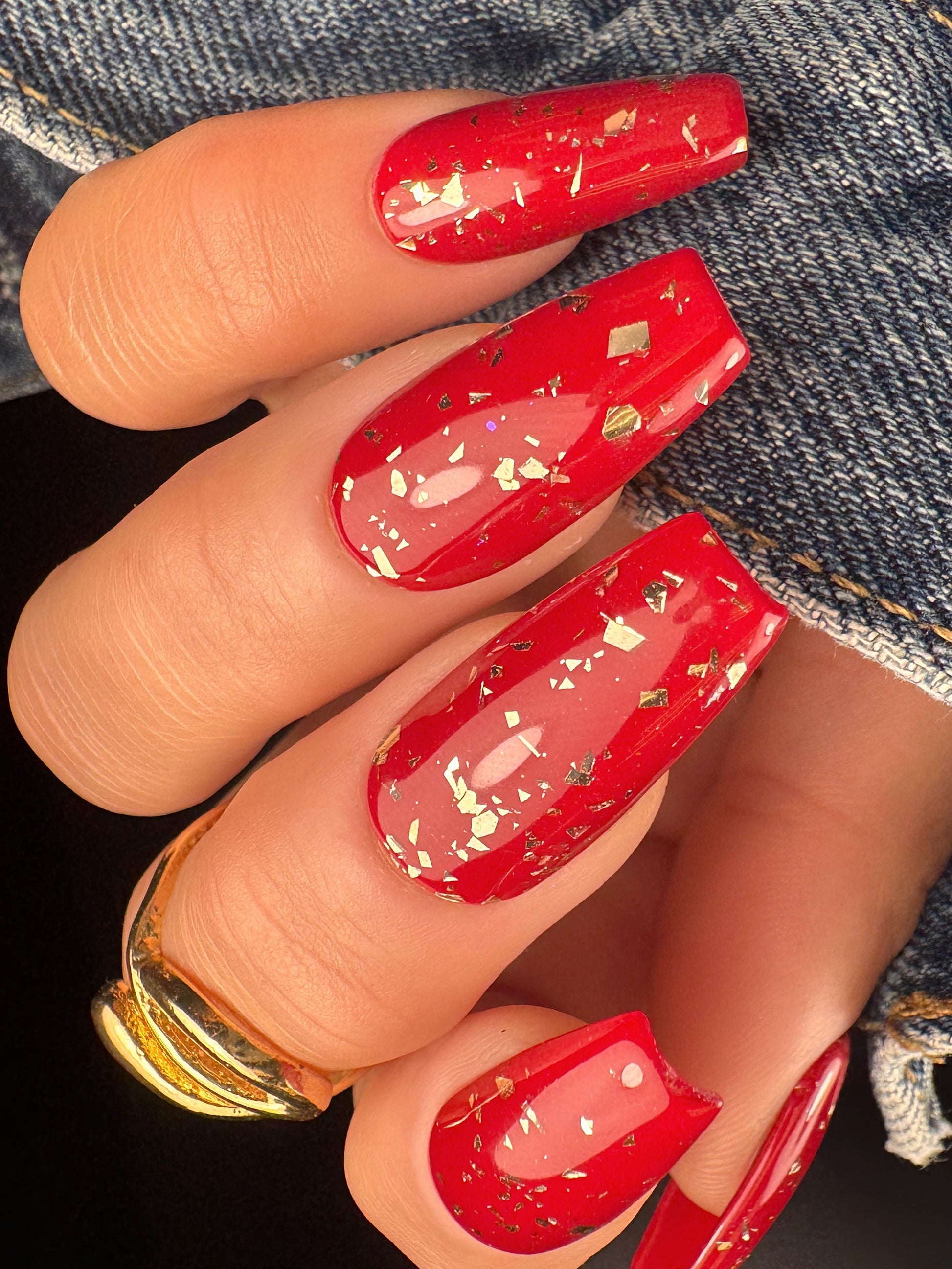 Red nails with gold glitter on a hand wearing a gold ring, against a denim background.