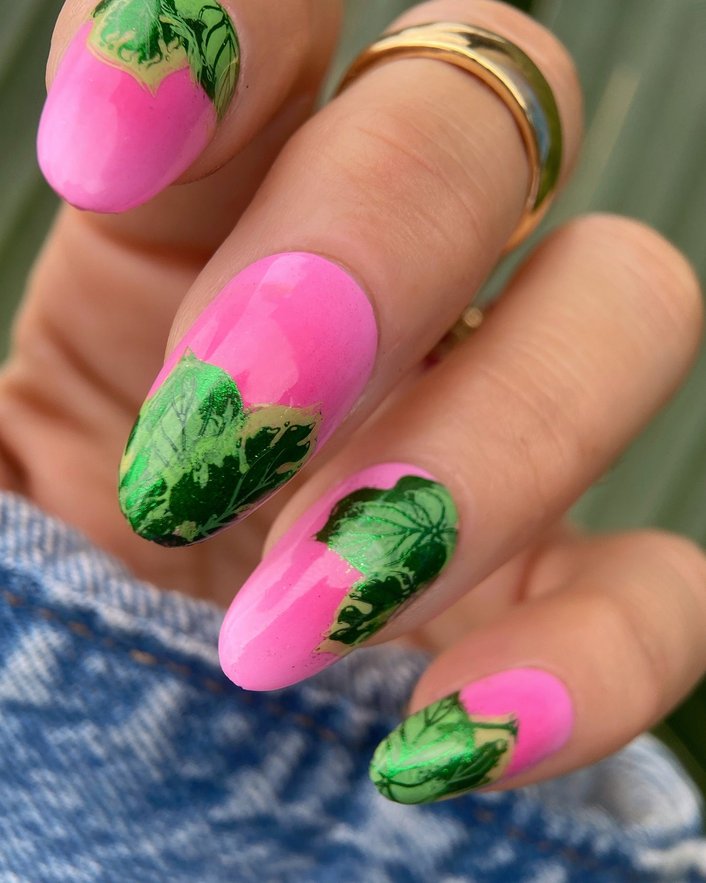 Nails with pink polish and green leaf designs, held by a person wearing a gold ring.