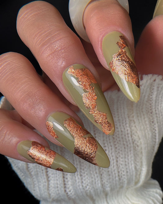 Nails with olive green polish and copper foil accents on a textured white fabric background.