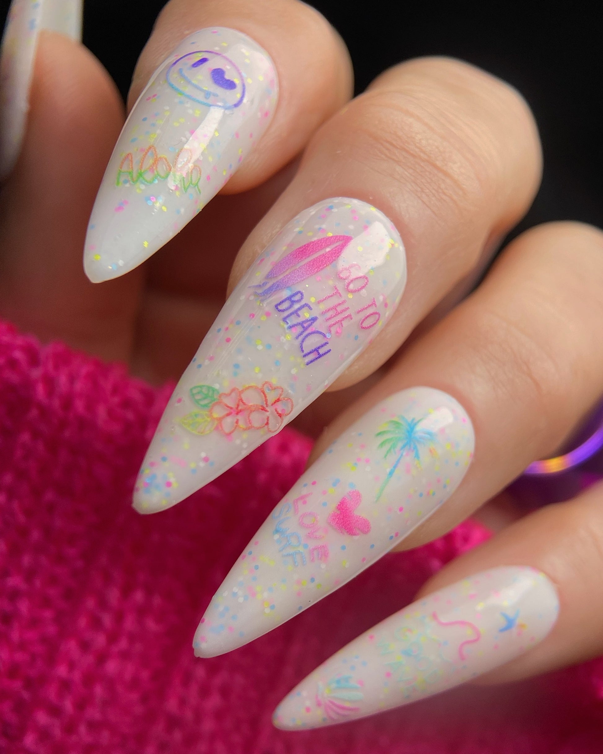 Nails with colorful designs and text held by a hand against a pink background