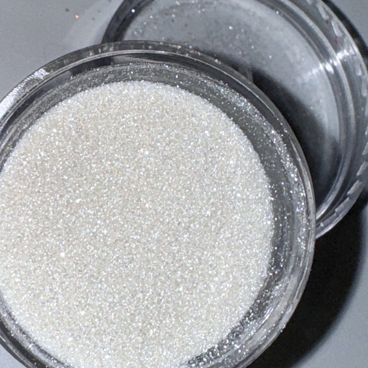 A close-up of a clear container filled with white fine glitter, intended for nail art.