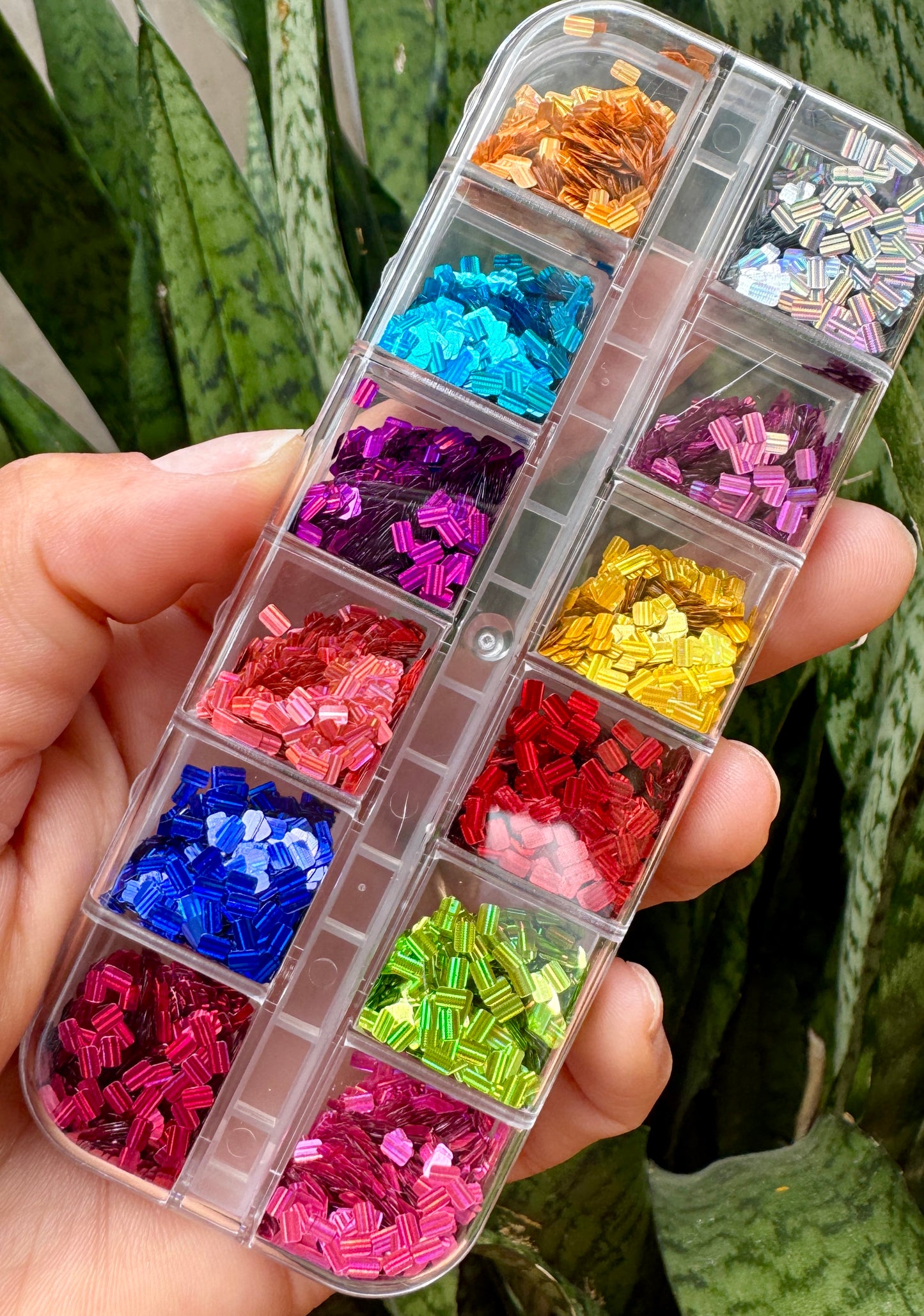 Transparent container with compartments filled with colorful rectangle nail glitter held by a hand against a green leafy background.