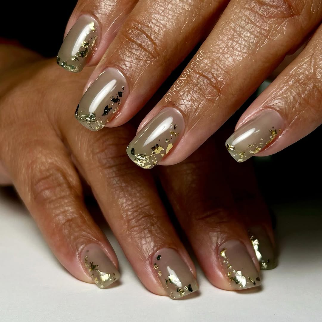A close-up image of a person's nails which are painted with a neutral color polish and adorned with gold flakes to create a metallic nail art design.
