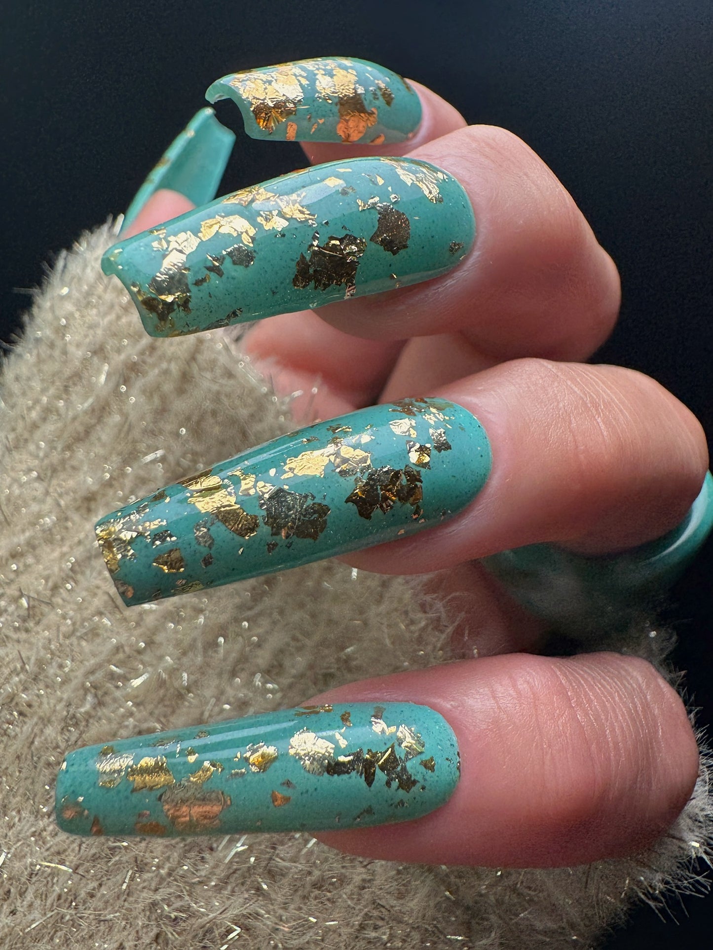 Turquoise nails with gold flakes on a hand against a dark background