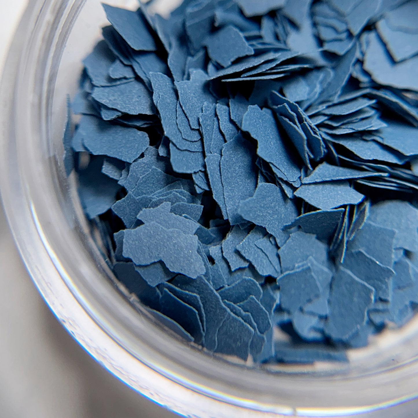 A close-up view of a jar containing blue matte flakes intended for nail art.