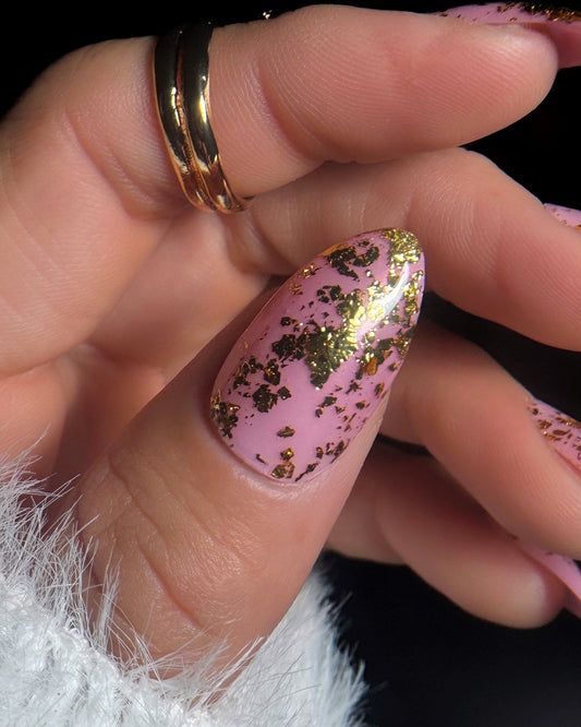 Close-up of a hand with pink nails and gold flake design, wearing a gold ring.