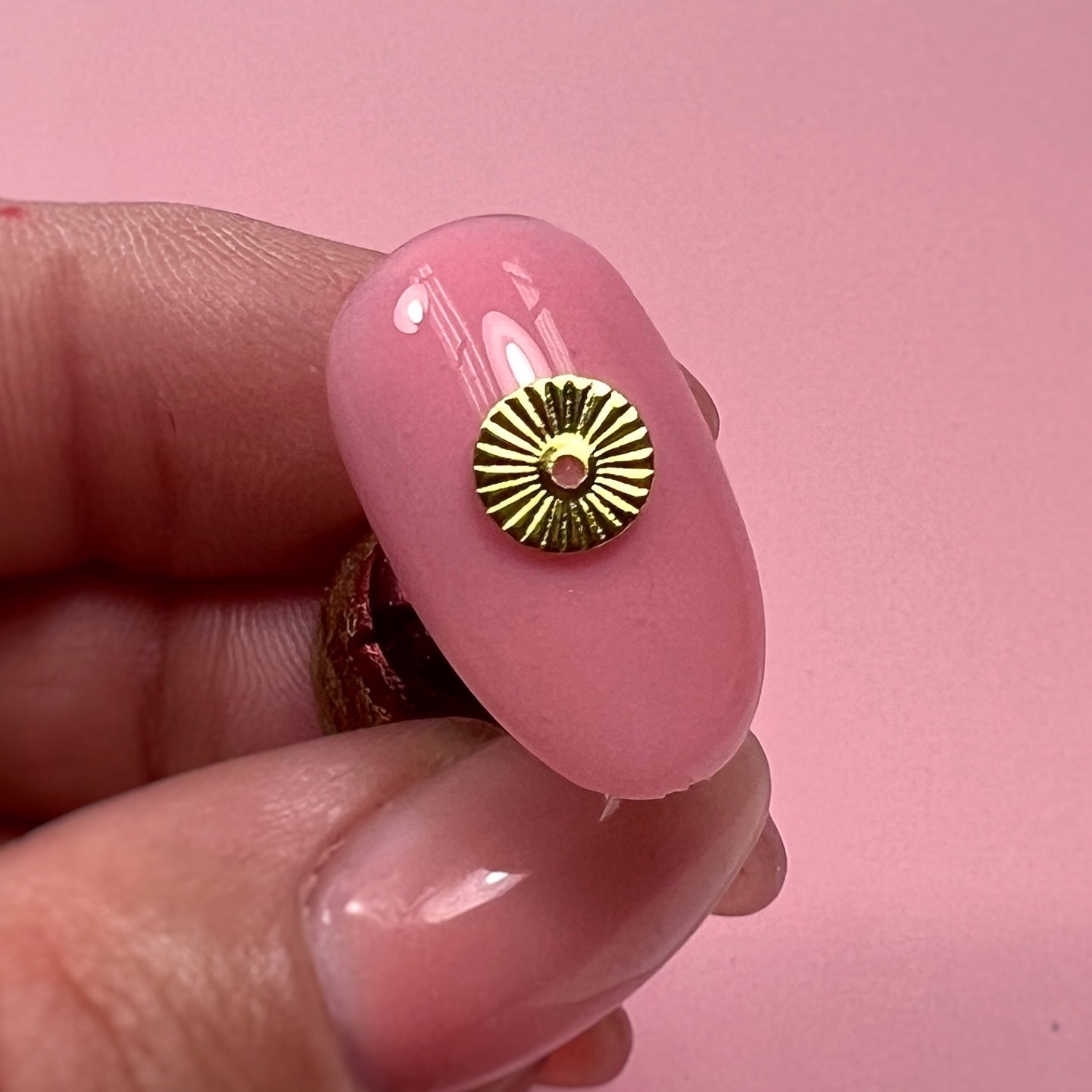 Pink nail tip with gold charm held between fingers against a pink background
