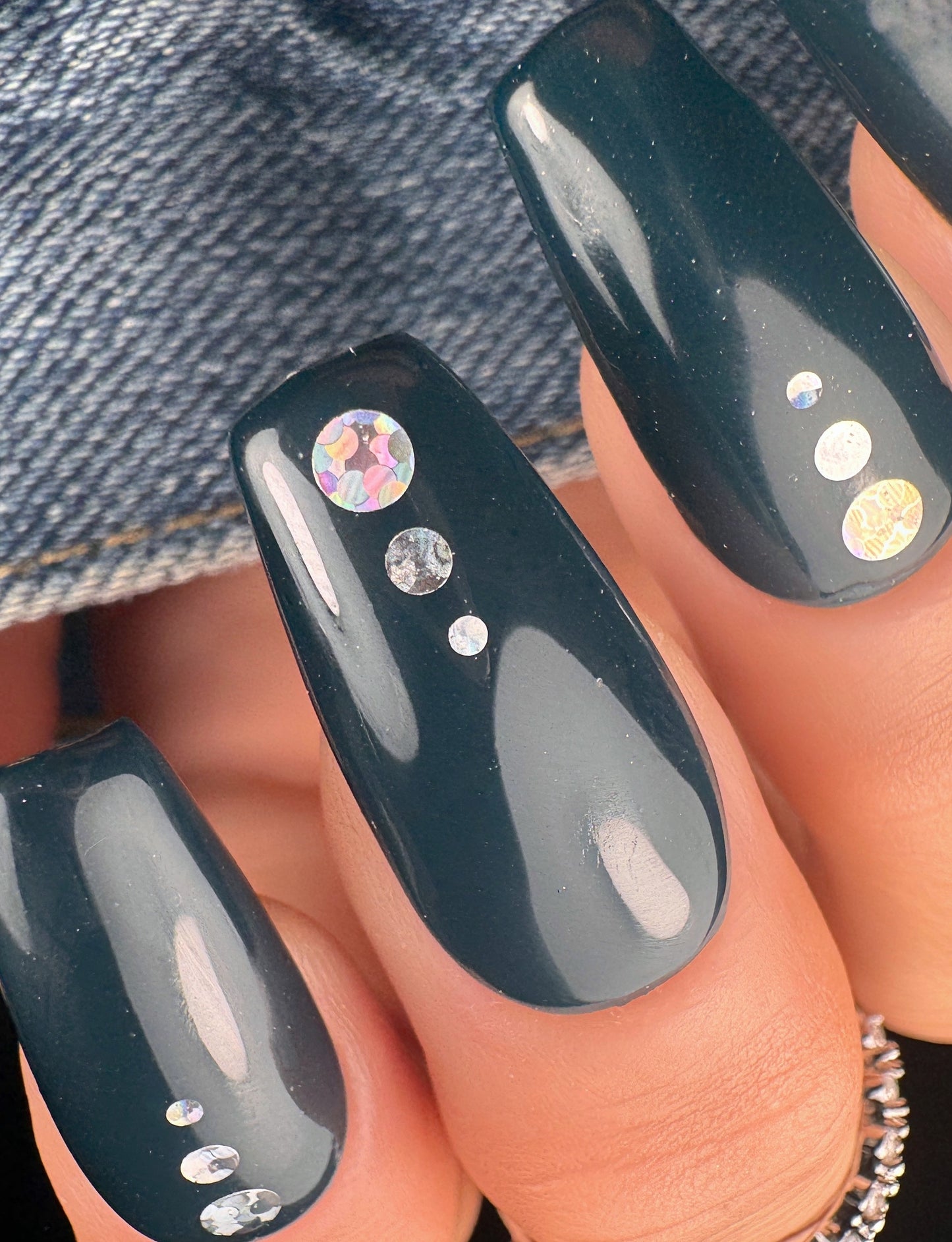 Black nail polish with encapsulated silver holographic glitter on a person's hand against a denim background