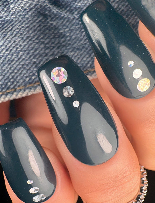 Black nail polish with encapsulated silver holographic glitter on a person's hand against a denim background