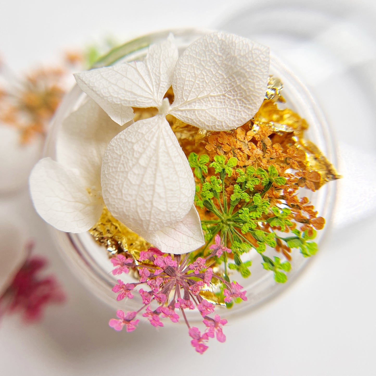 Close-up of a transparent container with white flowers, green leaves, and pink flowers on a blurred background.