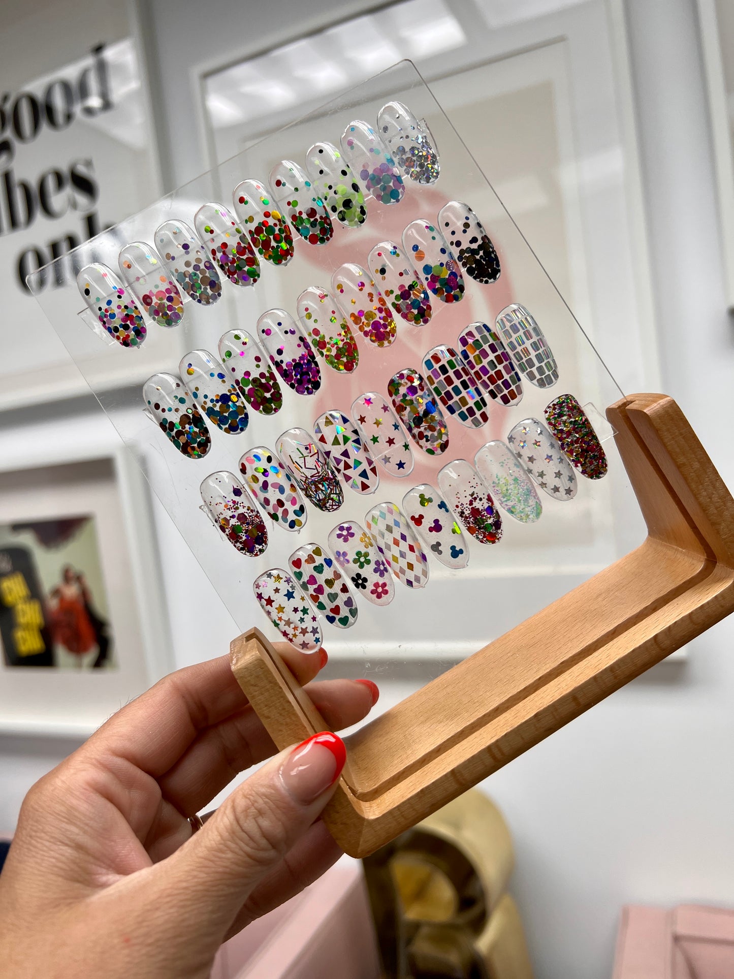 Hand Holding Wood and Clear Acrylic Nail Art Display with Sample Nail Art Tips, Against a White Wall with Picture Frames