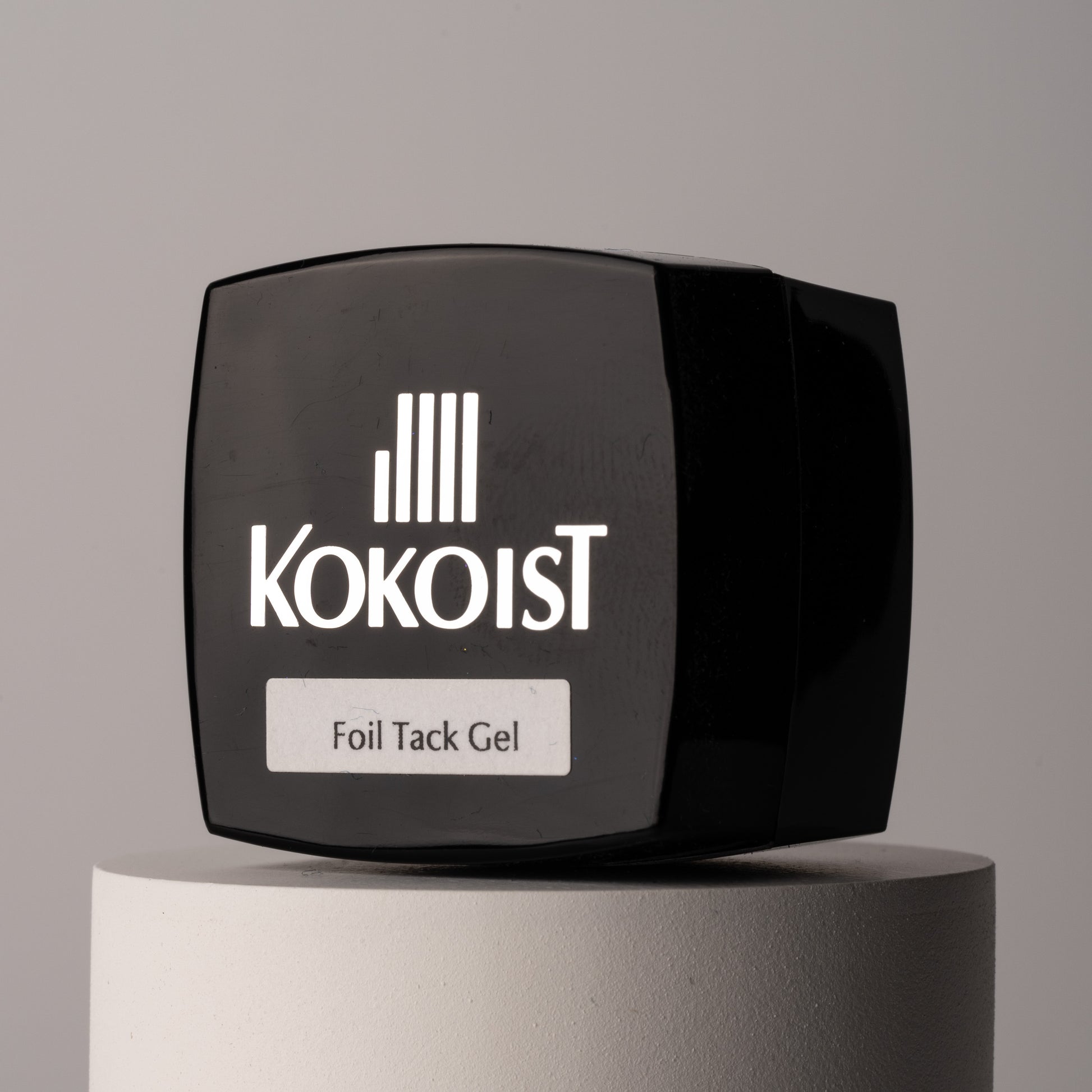 A black square bottle with the KOKOIST brand logo and the product name 'Foil Tack Gel' on it, placed on a grey cylindrical pedestal.