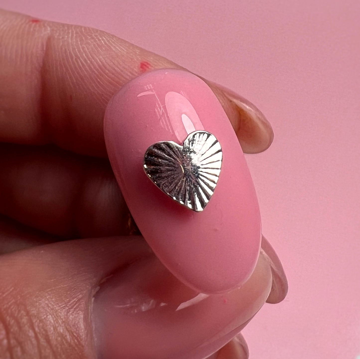 Pink nail with a silver heart-shaped charm held between fingers against a pink background