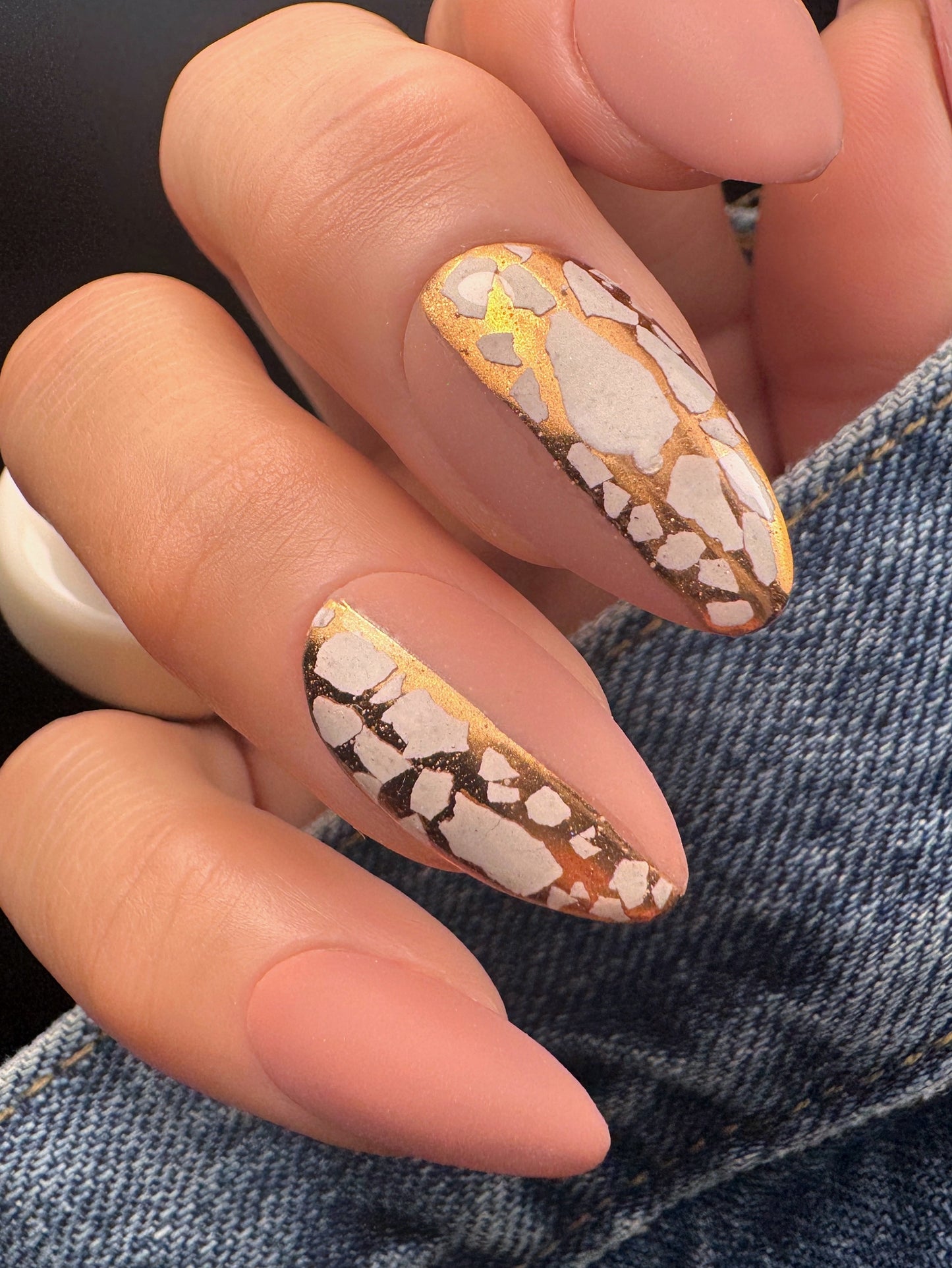 Close-up of a hand with almond-shaped nails featuring a mosaic design in white and gold, against a denim background.