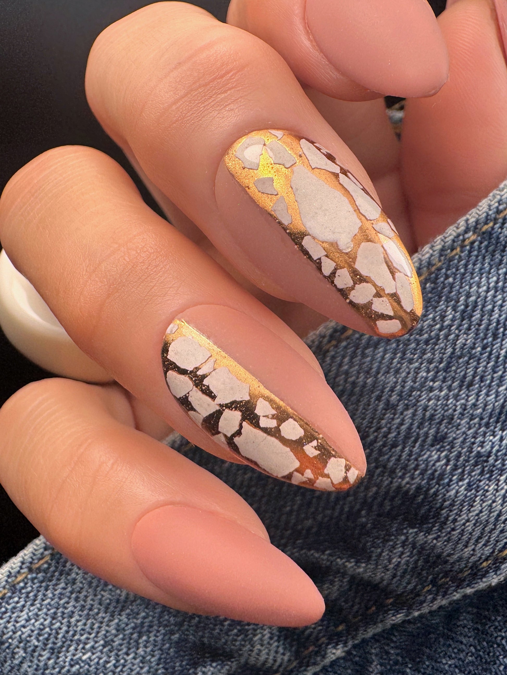 Close-up of a hand with almond-shaped nails featuring a mosaic design in white and gold, against a denim background.