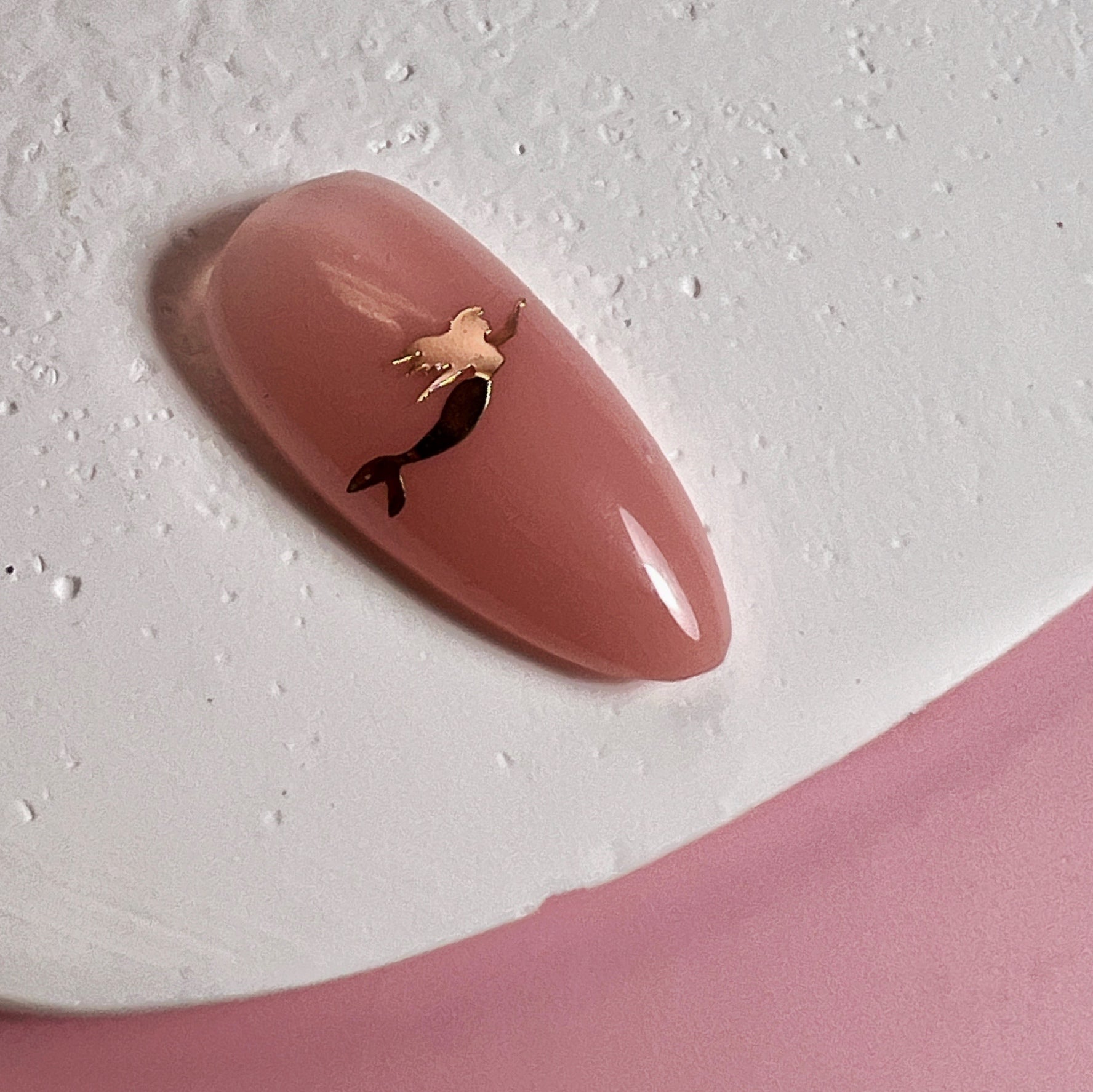 Oink nail art tip with gold mermaid charm on white textured surface on pink background.
