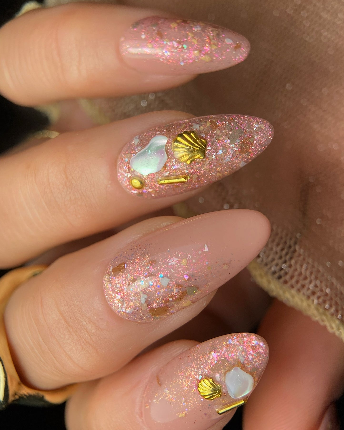 Close-up of a hand with glittery pink nails featuring gold shells and pearls.