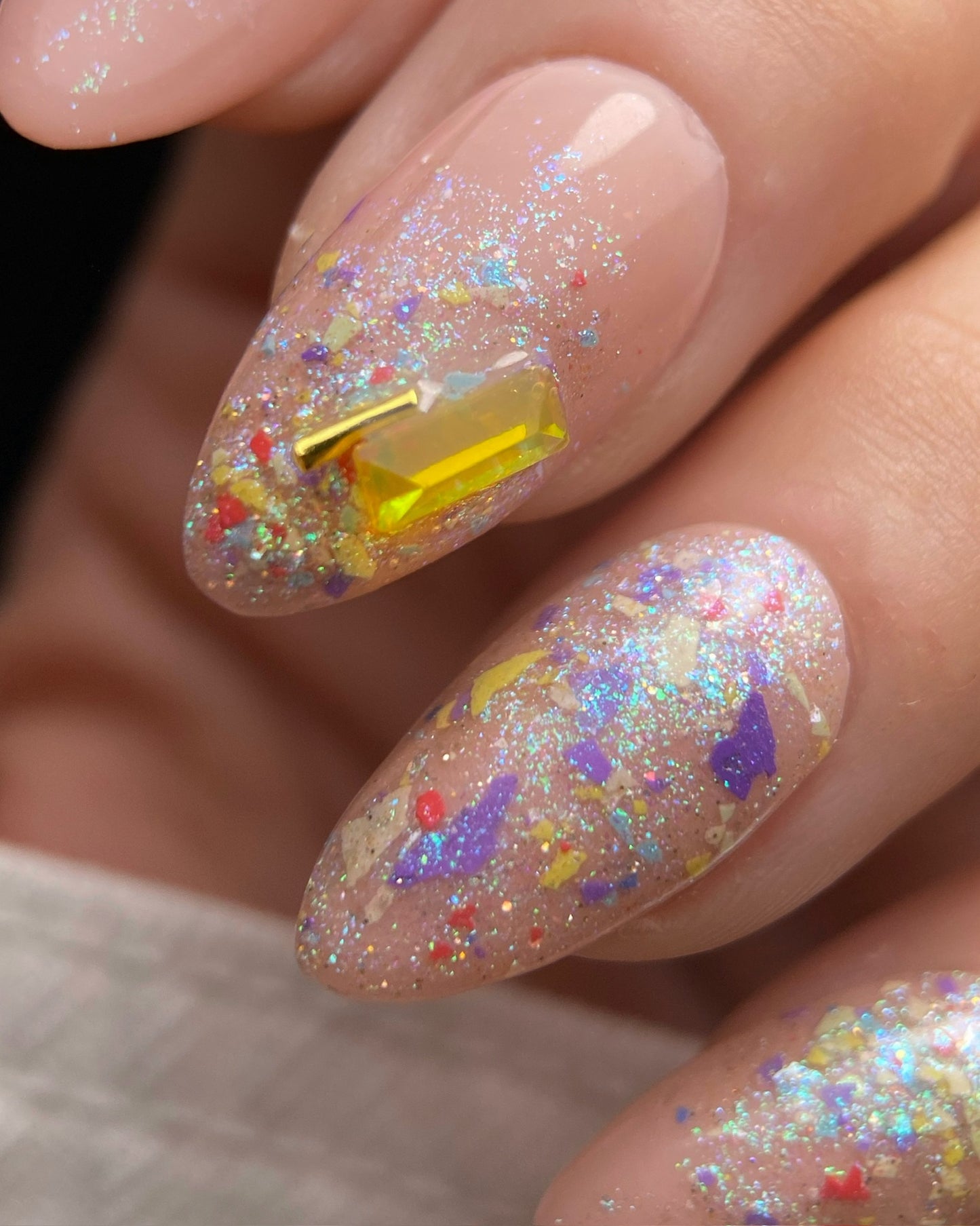 Nail art with colorful glitter and a yellow gemstone on a neutral background