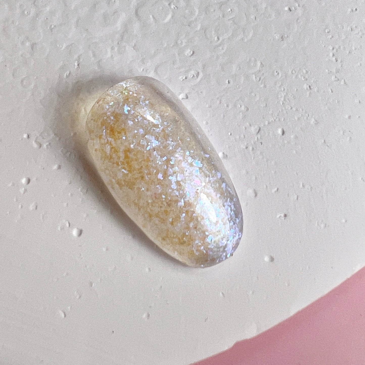 Close up of nail tip with glittery finish on textured white surface and pink background.