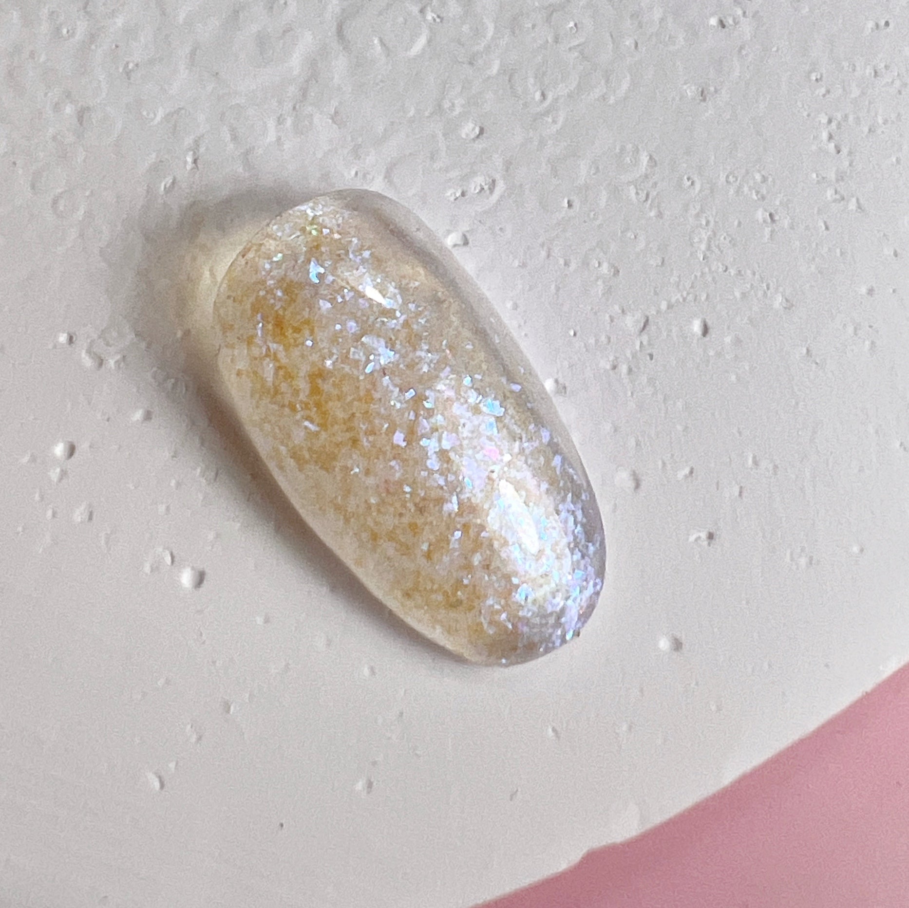 Close up of nail tip with glittery finish on textured white surface and pink background.