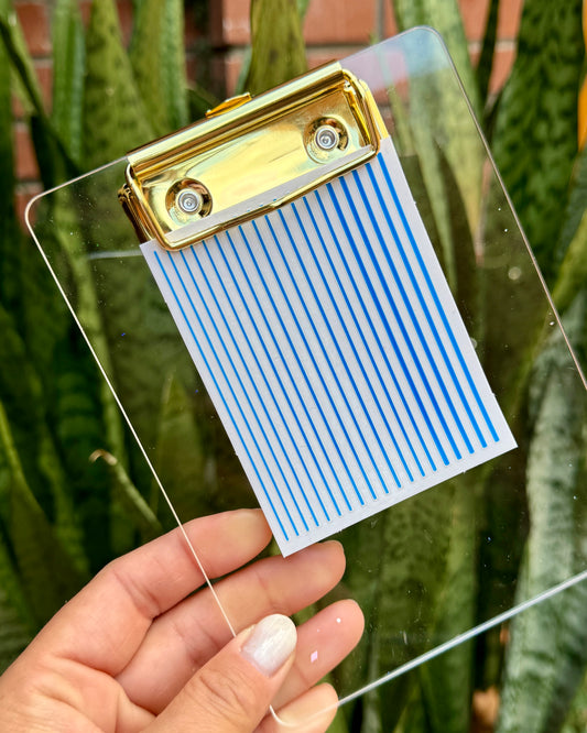 Clear clipboard with nail decal sheet and gold clip held by a hand in front of green plants