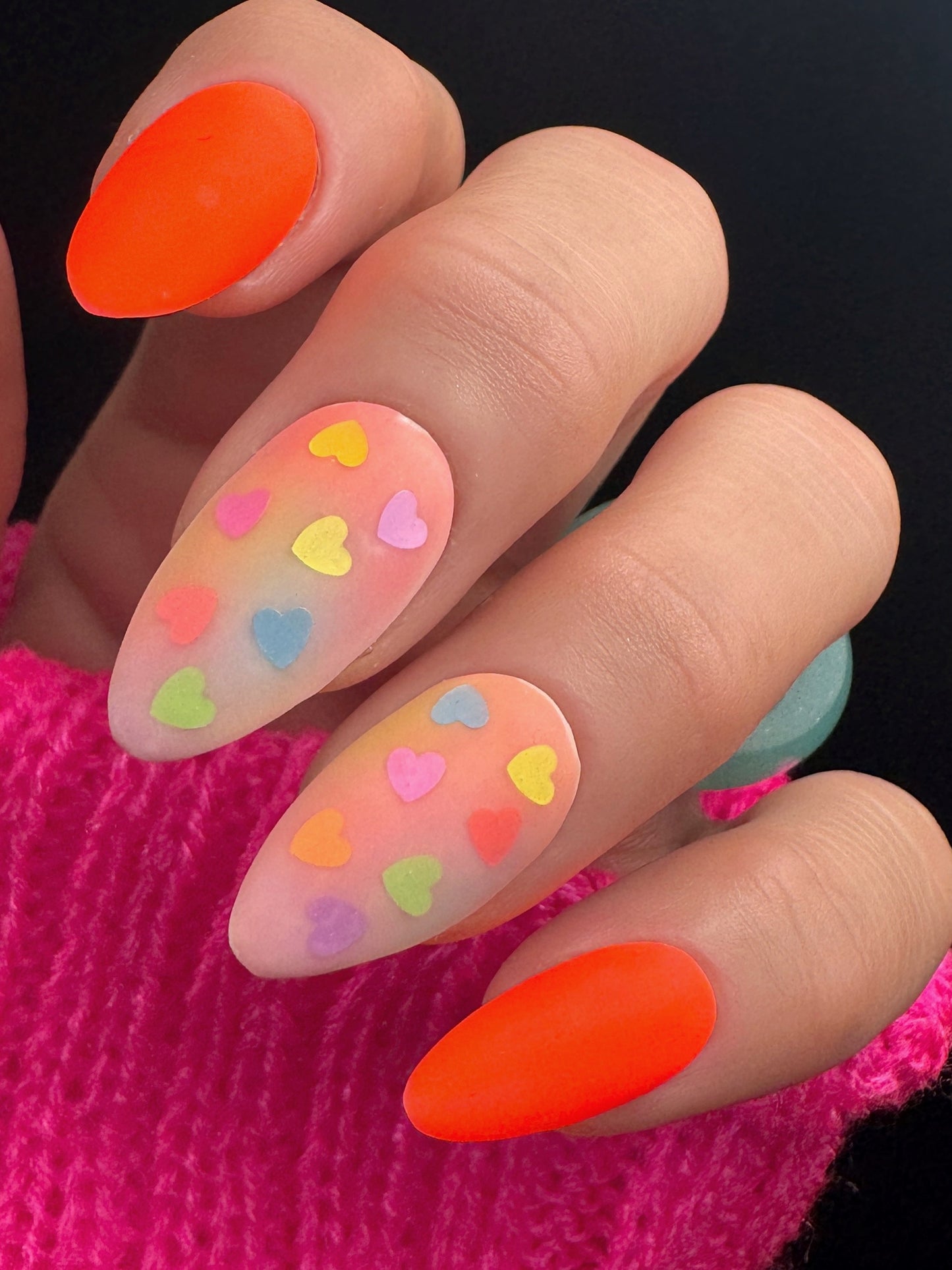 Nails with colorful heart designs and neon orange accents on a pink textured background