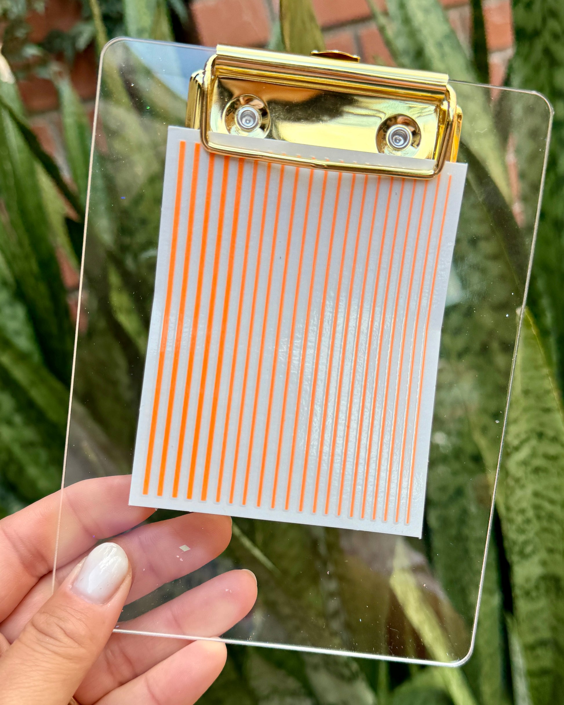 Clear clipboard with gold clip holding a nail decal sheet with orange stripes against a green plant background