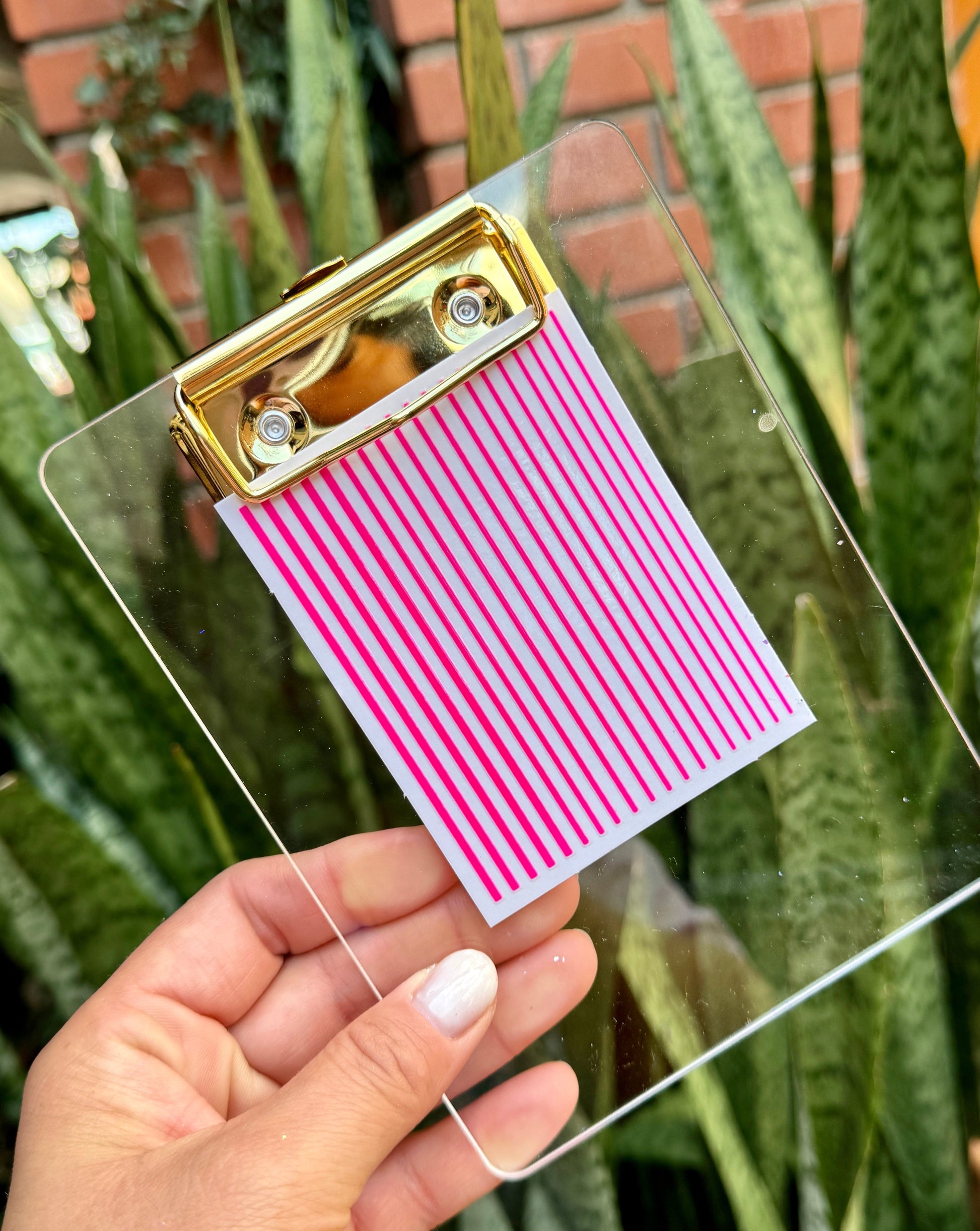Clear clipboard with gold clip and nail decal sheet held by a hand against a plant background