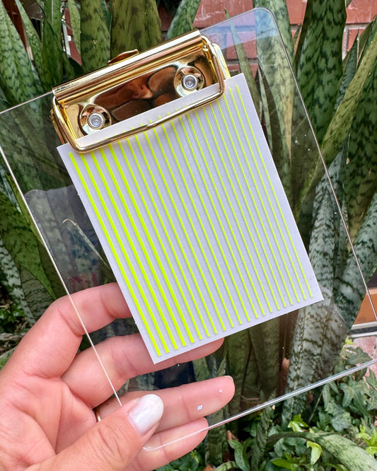 Clear clipboard with gold clip and nail decal sheet held by a hand against a plant background