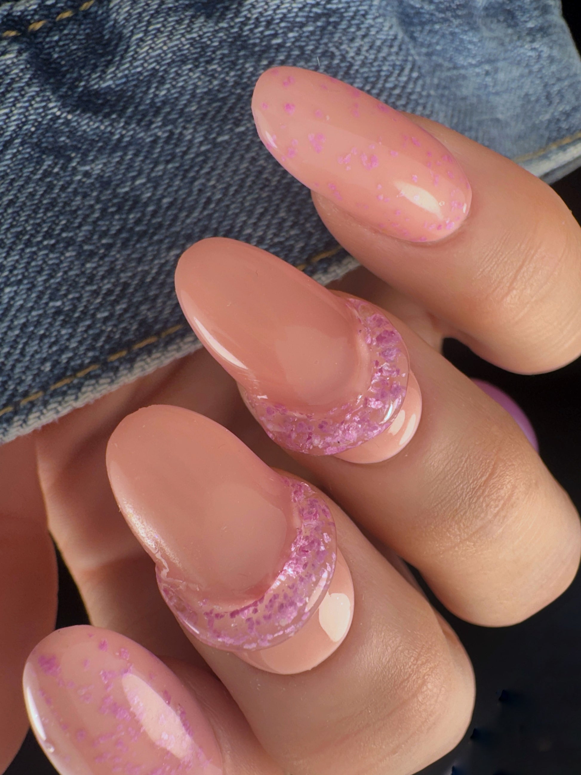 Close-up of a hand with matte speckled glitter and clear 3D art on neutral nail on a denim background