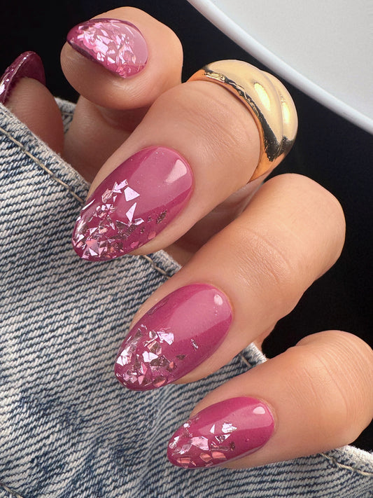 Hand with pink glitter nail art wearing a gold ring on denim fabric.