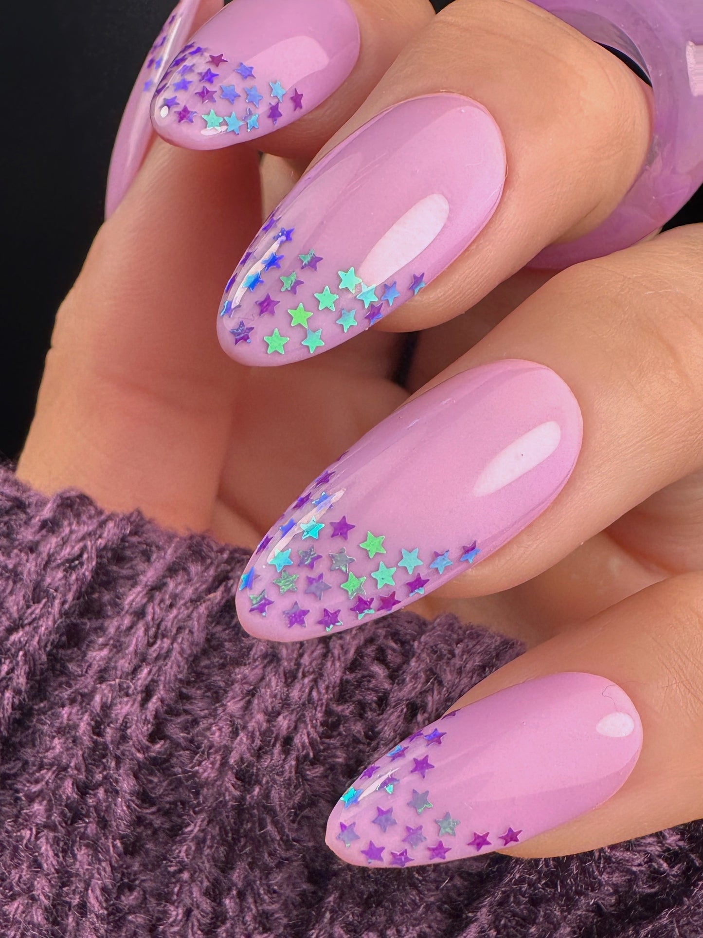 Hand with purple nails featuring purple star glitter design on a dark background
