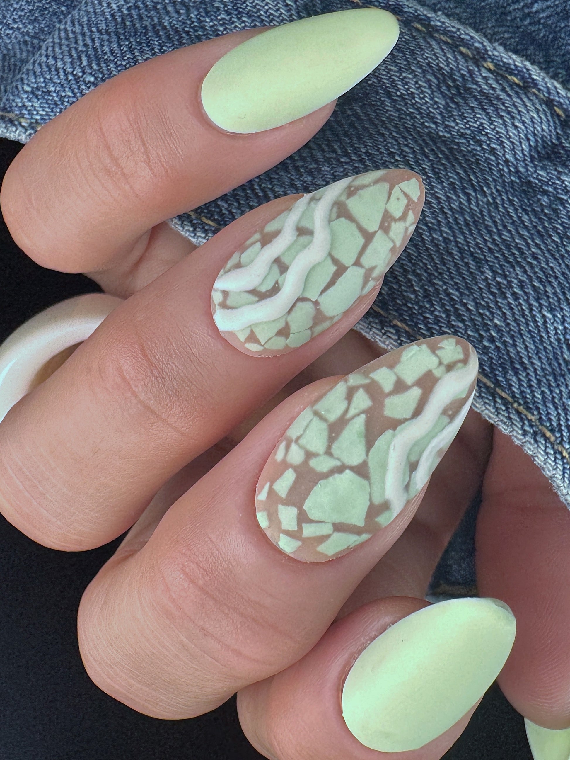 Close-up of a hand with light green nails featuring a mosaic pattern and white detail, against a denim background.